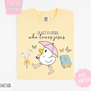Puede incluir: Una camiseta amarilla con el texto "Just a girl who loves Jesus" en negro. El dise&ntilde;o tambi&eacute;n incluye un pato de dibujos animados con botas de lluvia y sosteniendo un paraguas, junto con flores y mariposas.