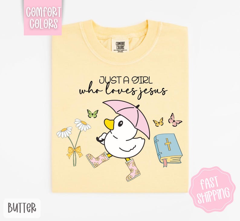 Puede incluir: Una camiseta amarilla con el texto "Just a girl who loves Jesus" en negro. El dise&ntilde;o tambi&eacute;n incluye un pato de dibujos animados con botas de lluvia y sosteniendo un paraguas, junto con flores y mariposas.