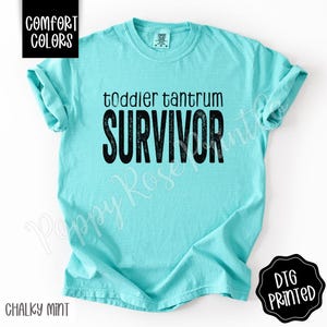 May include: A chalky mint-colored t-shirt with the words "toddler tantrum SURVIVOR" printed in black. The shirt has short sleeves and a crew neck. The words "Comfort Colors" and "DTG Printed" are also visible.