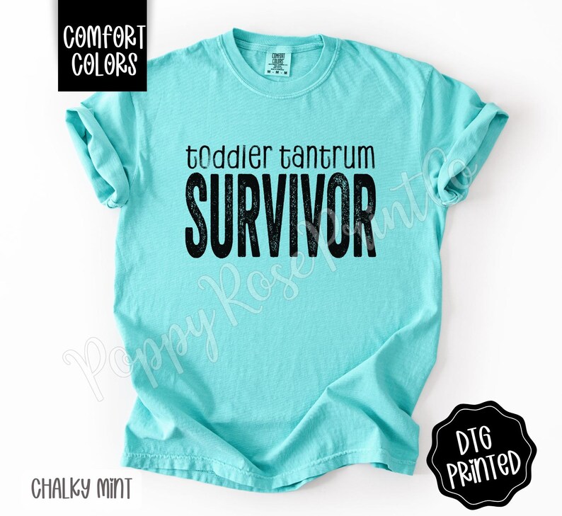 May include: A chalky mint-colored t-shirt with the words "toddler tantrum SURVIVOR" printed in black. The shirt has short sleeves and a crew neck. The words "Comfort Colors" and "DTG Printed" are also visible.
