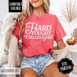 May include: A watermelon-colored t-shirt with the words "Hard Fought Hallelujah" in white script. The shirt is a Comfort Colors brand and is DTG printed. The model is wearing denim shorts and a cream-colored cardigan.