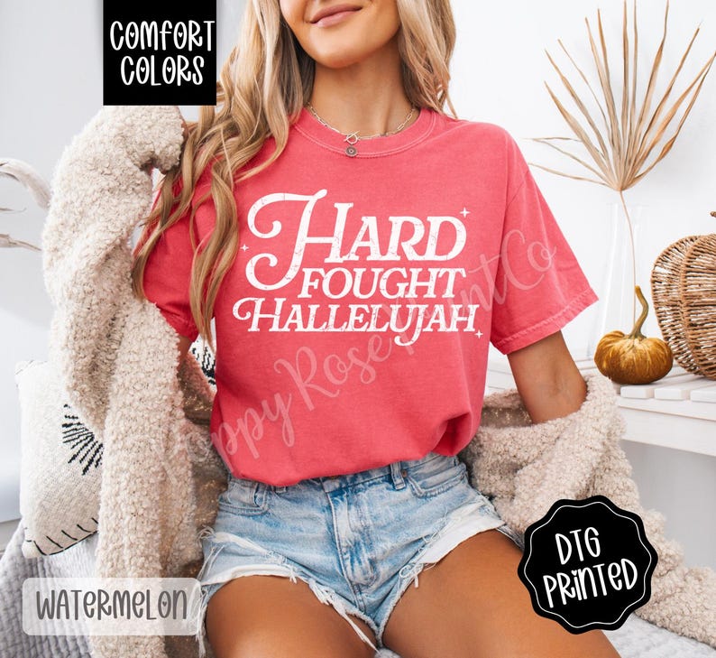 May include: A watermelon-colored t-shirt with the words "Hard Fought Hallelujah" in white script. The shirt is a Comfort Colors brand and is DTG printed. The model is wearing denim shorts and a cream-colored cardigan.