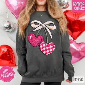 May include: Dark grey sweatshirt featuring a cherry design with a white bow and two heart-shaped cherries. One cherry is solid pink, and the other has a pink and white checkered pattern. The sweatshirt has the words "Comfort Colors" and "Pepper" printed on it.