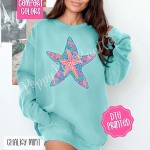 May include: A light blue sweatshirt with a colorful starfish design. The sweatshirt has a crew neck and long sleeves. The text "Comfort Colors" is printed on the top of the sweatshirt. The text "DTG Printed" is printed on a pink circle on the bottom right of the sweatshirt. The text "Chalky Mint" is printed on the bottom left of the sweatshirt.