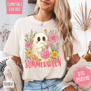 May include: Ivory-colored t-shirt featuring a cartoon ghost enjoying a pink drink with watermelon slices, surrounded by flowers. The text "SUMMERWEEN" is in pink. The shirt has "COMFORT COLORS" and "DTG PRINTED" labels.