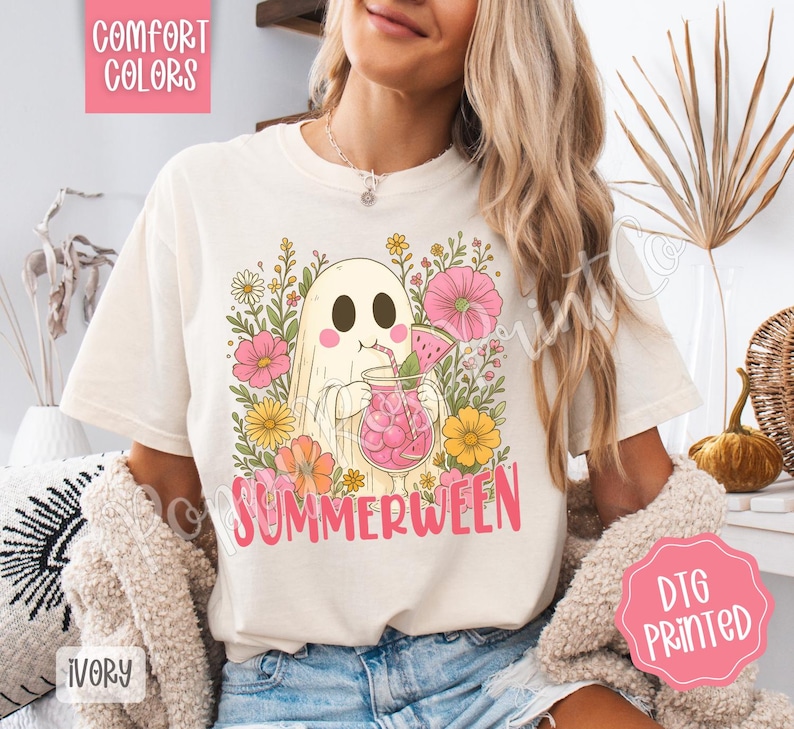 May include: Ivory-colored t-shirt featuring a cartoon ghost enjoying a pink drink with watermelon slices, surrounded by flowers. The text "SUMMERWEEN" is in pink. The shirt has "COMFORT COLORS" and "DTG PRINTED" labels.