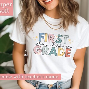 Op de afbeelding: Wit T-shirt met de tekst "FIRST" in roze, "Mrs. Miller" in blauw en "GRADE" in geel. De tekst is zwart omrand en heeft een madeliefje-ontwerp. Het shirt is te personaliseren met de naam van een leraar.