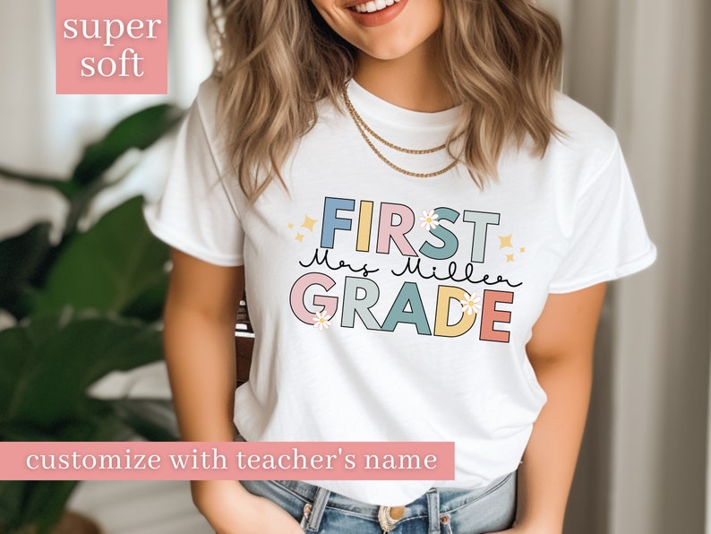 Op de afbeelding: Wit T-shirt met de tekst "FIRST" in roze, "Mrs. Miller" in blauw en "GRADE" in geel. De tekst is zwart omrand en heeft een madeliefje-ontwerp. Het shirt is te personaliseren met de naam van een leraar.