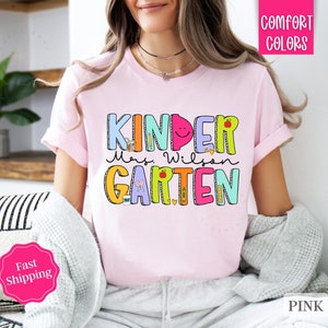May include: Pink t-shirt with the text "Kinder Mrs. Wilson Garten" in colorful letters with illustrations of school supplies and a smiling face.