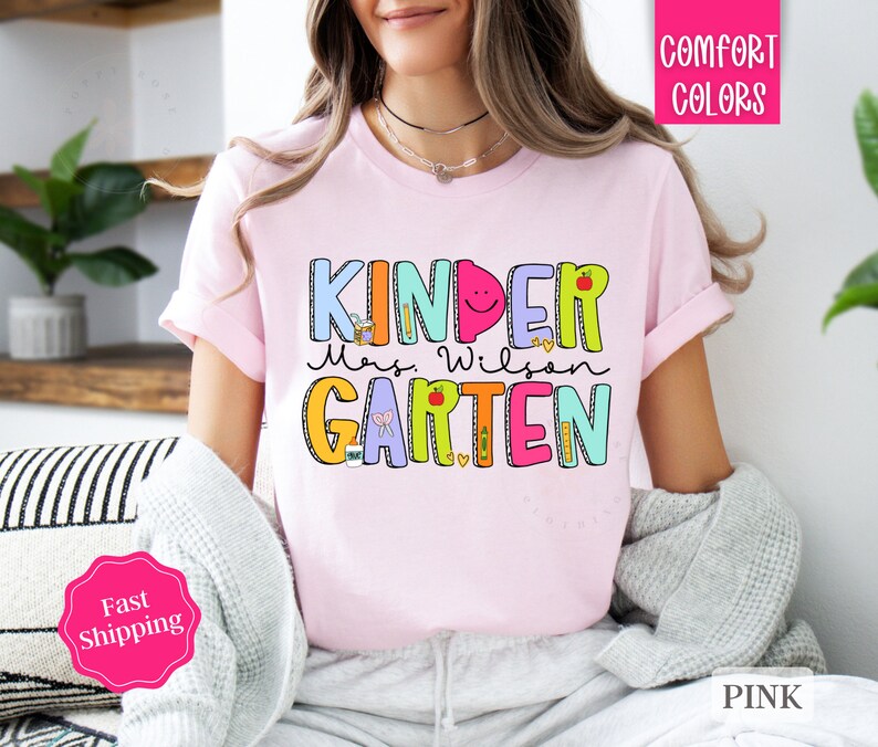 May include: Pink t-shirt with the text "Kinder Mrs. Wilson Garten" in colorful letters with illustrations of school supplies and a smiling face.