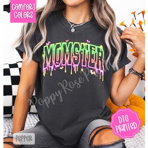 May include: Dark gray t-shirt with the word "MOMSTER" in neon green, pink, and purple dripping font. The shirt has short sleeves and is labeled "Comfort Colors" and "DTG Printed".