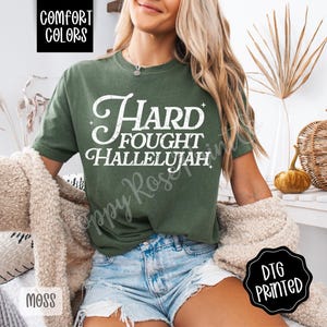 May include: Moss green t-shirt with the words "Hard Fought Hallelujah" in white script. The shirt is part of the Comfort Colors collection and is DTG printed. The model is wearing blue jean shorts.