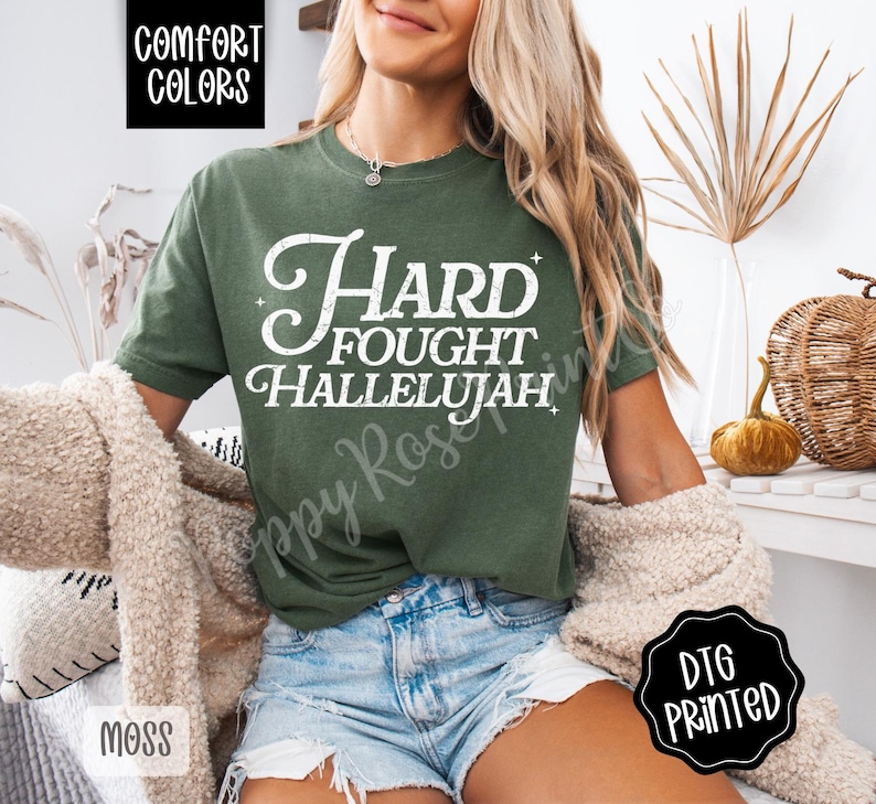 May include: Moss green t-shirt with the words "Hard Fought Hallelujah" in white script. The shirt is part of the Comfort Colors collection and is DTG printed. The model is wearing blue jean shorts.