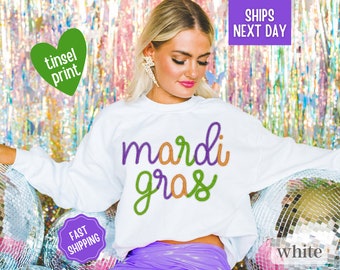 May include: White sweatshirt with the text "Mardi Gras" in green, purple and orange embroidery. The sweatshirt is on a model wearing a purple skirt.