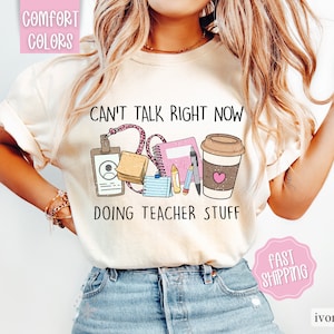 May include: A white t-shirt with the text "Can't talk right now doing teacher stuff" and a graphic of teacher supplies, including a coffee cup, a notebook, pencils, and a name tag. The shirt is on a model wearing blue jeans.