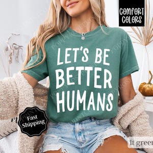 May include: A teal green t-shirt with the text "Let's Be Better Humans" printed in white. The shirt is made of a soft, comfortable fabric and has a relaxed fit.