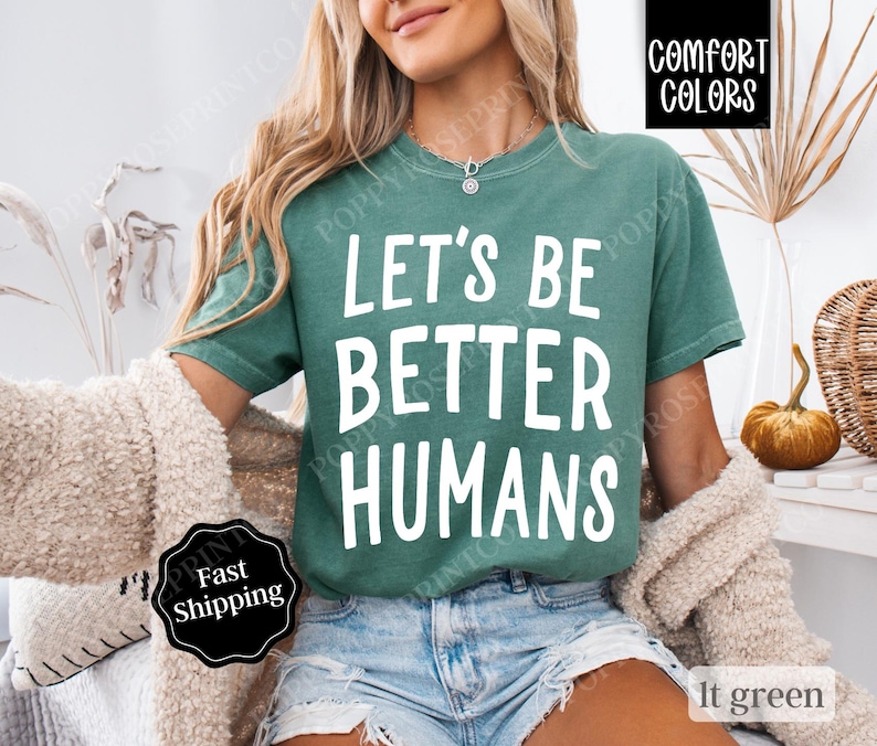 May include: A teal green t-shirt with the text "Let's Be Better Humans" printed in white. The shirt is made of a soft, comfortable fabric and has a relaxed fit.