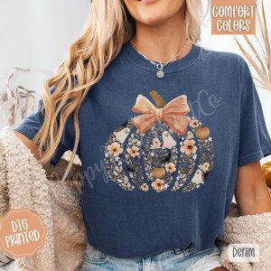 Pu&ograve; includere: Una maglietta blu navy con un design a zucca pieno di fiori, fantasmi e un fiocco. Il testo "Comfort Colors" &egrave; nell'angolo in alto a destra e "DTG Printed" nell'angolo in basso a sinistra. La parola "Denim" &egrave; nell'angolo in basso a destra.