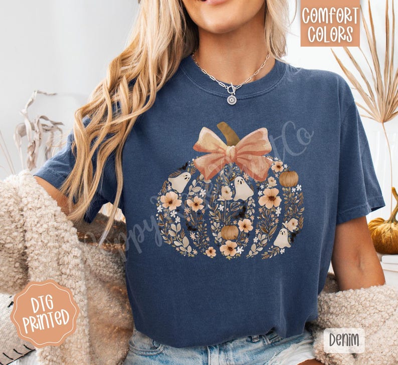Pu&ograve; includere: Una maglietta blu navy con un design a zucca pieno di fiori, fantasmi e un fiocco. Il testo "Comfort Colors" &egrave; nell'angolo in alto a destra e "DTG Printed" nell'angolo in basso a sinistra. La parola "Denim" &egrave; nell'angolo in basso a destra.