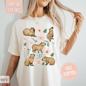 Pu&ograve; includere: Una maglietta bianca con un disegno di capibara e fiori rosa. Il testo "Comfort Colors" &egrave; nell'angolo in alto a sinistra dell'immagine.