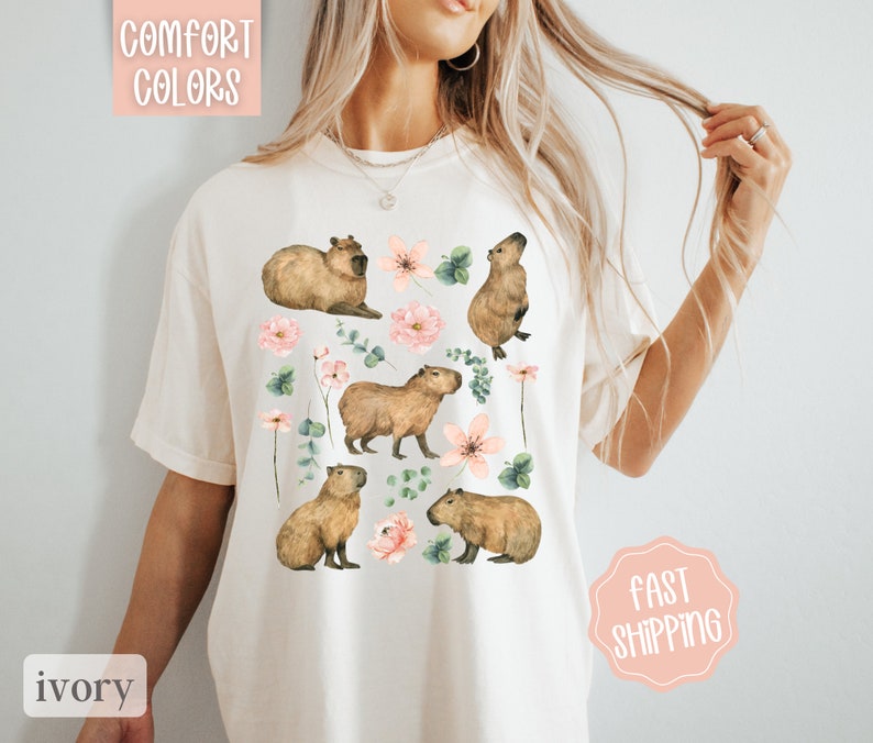 Pu&ograve; includere: Una maglietta bianca con un disegno di capibara e fiori rosa. Il testo "Comfort Colors" &egrave; nell'angolo in alto a sinistra dell'immagine.