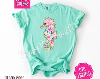 May include: A light turquoise t-shirt featuring a colorful seahorse design. The seahorse is decorated with pink, yellow, and blue floral patterns and gold glitter accents. The shirt has the text "Comfort Colors", "Island Reef", and "DTG Printed".