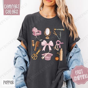 May include: A dark gray t-shirt with a graphic design of various hair salon tools, including a hair dryer, scissors, a hairbrush, a hair dye bowl, and a pink ribbon. The design is in a vintage style with a pink and gold color scheme.