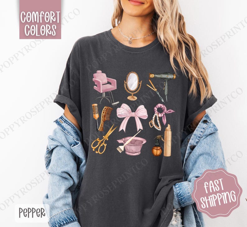 May include: A dark gray t-shirt with a graphic design of various hair salon tools, including a hair dryer, scissors, a hairbrush, a hair dye bowl, and a pink ribbon. The design is in a vintage style with a pink and gold color scheme.
