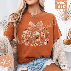 Pu&ograve; includere: T-shirt arancione con un design a zucca fatto di fiori, fantasmi e pipistrelli. La zucca ha un grande fiocco in cima. Le parole "Comfort Colors" sono stampate in alto a destra. Le parole "DTG Printed" sono stampate in basso a sinistra.