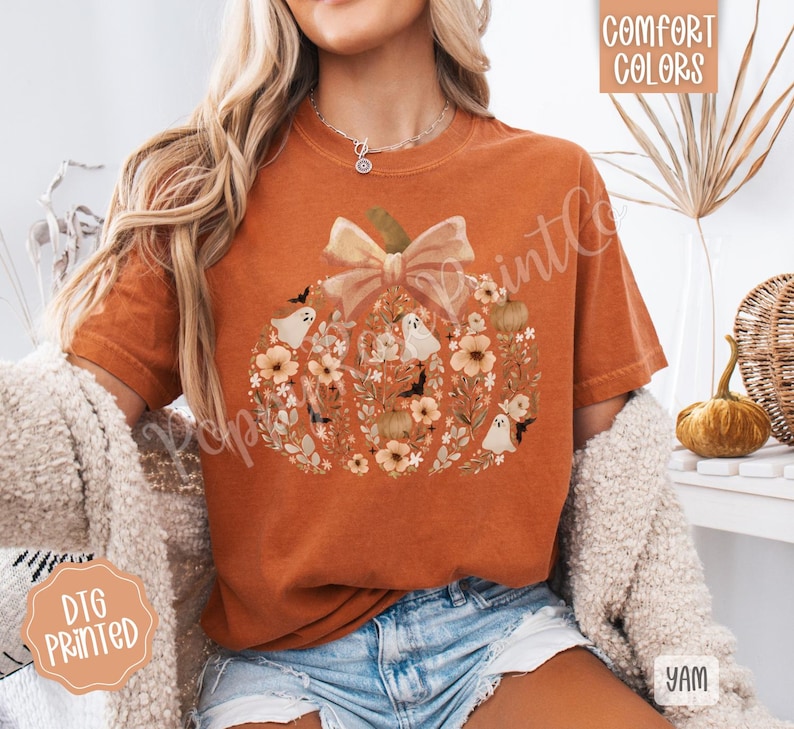 Pu&ograve; includere: T-shirt arancione con un design a zucca fatto di fiori, fantasmi e pipistrelli. La zucca ha un grande fiocco in cima. Le parole "Comfort Colors" sono stampate in alto a destra. Le parole "DTG Printed" sono stampate in basso a sinistra.