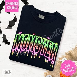 May include: Black t-shirt with the word "MONSTER" in neon green, pink, and yellow. The word has a dripping effect and is decorated with bats. The shirt is labeled "Comfort Colors" and "DTG Printed".