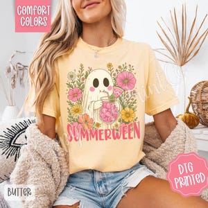 May include: Yellow t-shirt with a graphic of a ghost holding a pink drink, surrounded by flowers. The word "SUMMERWEEN" is printed in pink. The shirt has the words "COMFORT COLORS" and "BUTTER" printed on it.