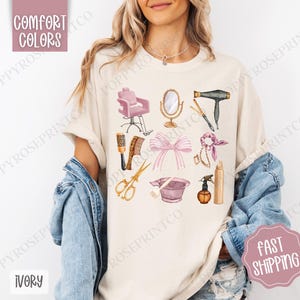 May include: A beige t-shirt with a design of various hair salon tools, including a pink salon chair, a hairdryer, scissors, combs, a hairspray bottle, and a pink hair dye bowl. The design is in a watercolor style with gold accents.