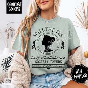 Pu&ograve; includere: T-shirt verde chiaro con la scritta "SPILL THE TEA" sopra una silhouette di profilo femminile. Sotto, si legge "Lady Whistledown's SOCIETY PAPERS". La maglietta ha anche le parole "COMFORT COLORS" e "DTG PRINTED".