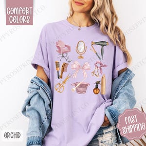 May include: A purple t-shirt with a design of various hair salon tools, including a hair dryer, scissors, a hairbrush, a styling chair, a hairspray bottle, and a hair tie. The design is in a watercolor style with pink and gold accents.