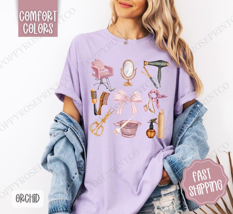 May include: A purple t-shirt with a design of various hair salon tools, including a hair dryer, scissors, a hairbrush, a styling chair, a hairspray bottle, and a hair tie. The design is in a watercolor style with pink and gold accents.