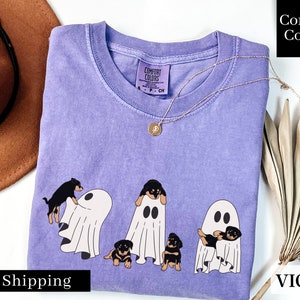May include: A purple t-shirt with a graphic of four black dogs dressed as ghosts. The text "Comfort Colors" is on the shirt. The shirt is laid flat on a white surface.