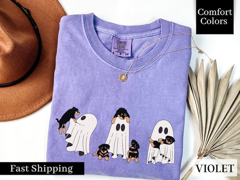May include: A purple t-shirt with a graphic of four black dogs dressed as ghosts. The text "Comfort Colors" is on the shirt. The shirt is laid flat on a white surface.