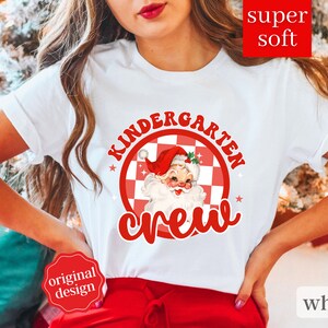 May include: White t-shirt with a red and white checkered circle design featuring a cartoon Santa Claus with the text "Kindergarten Crew".