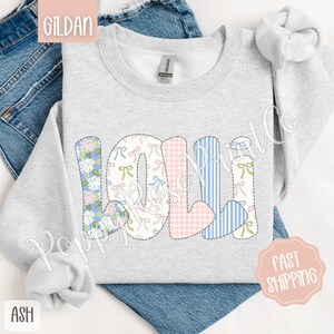 May include: A light gray sweatshirt with the word "LOLI" spelled out in colorful patchwork letters. The letters are made of different fabrics with floral, plaid, and striped patterns. The sweatshirt is a Gildan brand.