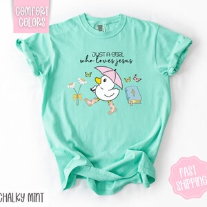 Puede incluir: Una camiseta azul turquesa con un gr&aacute;fico de un pato con botas de lluvia y sosteniendo un paraguas. El texto "Just a girl who loves Jesus" est&aacute; impreso en la camiseta.