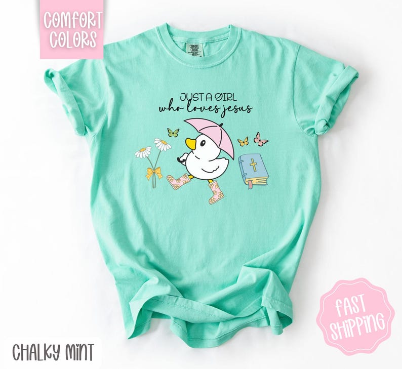 Puede incluir: Una camiseta azul turquesa con un gr&aacute;fico de un pato con botas de lluvia y sosteniendo un paraguas. El texto "Just a girl who loves Jesus" est&aacute; impreso en la camiseta.