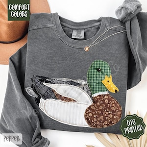 May include: A dark grey sweatshirt with a duck applique. The duck has a green checked head, a yellow beak, and a brown floral patterned body. The sweatshirt has the words "Comfort Colors" printed on a green label.