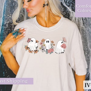 Peut inclure: Un t-shirt blanc avec un motif de trois fant&ocirc;mes tenant des fleurs. Les fant&ocirc;mes sont blancs avec des yeux et une bouche noirs. Les fleurs sont roses, orange et jaunes. Le texte "Comfort Colors" est dans le coin sup&eacute;rieur droit de l'image. Le texte "IVORY" est dans le coin inf&eacute;rieur droit de l'image.
