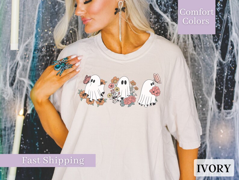 Peut inclure: Un t-shirt blanc avec un motif de trois fant&ocirc;mes tenant des fleurs. Les fant&ocirc;mes sont blancs avec des yeux et une bouche noirs. Les fleurs sont roses, orange et jaunes. Le texte "Comfort Colors" est dans le coin sup&eacute;rieur droit de l'image. Le texte "IVORY" est dans le coin inf&eacute;rieur droit de l'image.