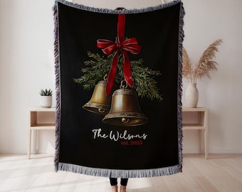 May include: Black woven throw blanket featuring a festive design with two golden bells, a red ribbon, and evergreen branches. The text "The Wilsons est. 2023" is displayed in white. The blanket has a fringed edge.