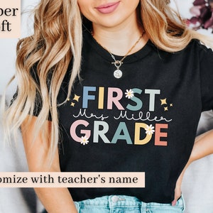 Op de afbeelding: Zwart T-shirt met de tekst "FIRST GRADE" in kleurrijke letters met bloemen en glitteraccenten. De tekst "Mrs. Miller" staat eronder in cursief. Het shirt is te personaliseren met de naam van een leraar.