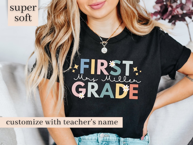 Op de afbeelding: Zwart T-shirt met de tekst "FIRST GRADE" in kleurrijke letters met bloemen en glitteraccenten. De tekst "Mrs. Miller" staat eronder in cursief. Het shirt is te personaliseren met de naam van een leraar.