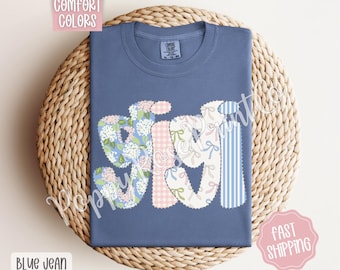 May include: A blue jean t-shirt with a patchwork design that spells out "Gigi". The letters are made of different fabrics and patterns, including floral, gingham, and stripes.