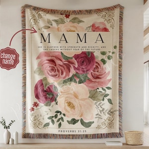 May include: A beige throw blanket with a floral design featuring pink and burgundy roses. The word "MAMA" is at the top, with the text "SHE IS CLOTHED WITH STRENGTH AND DIGNITY, AND SHE LAUGHS WITHOUT FEAR OF THE FUTURE. PROVERBS 31:25".